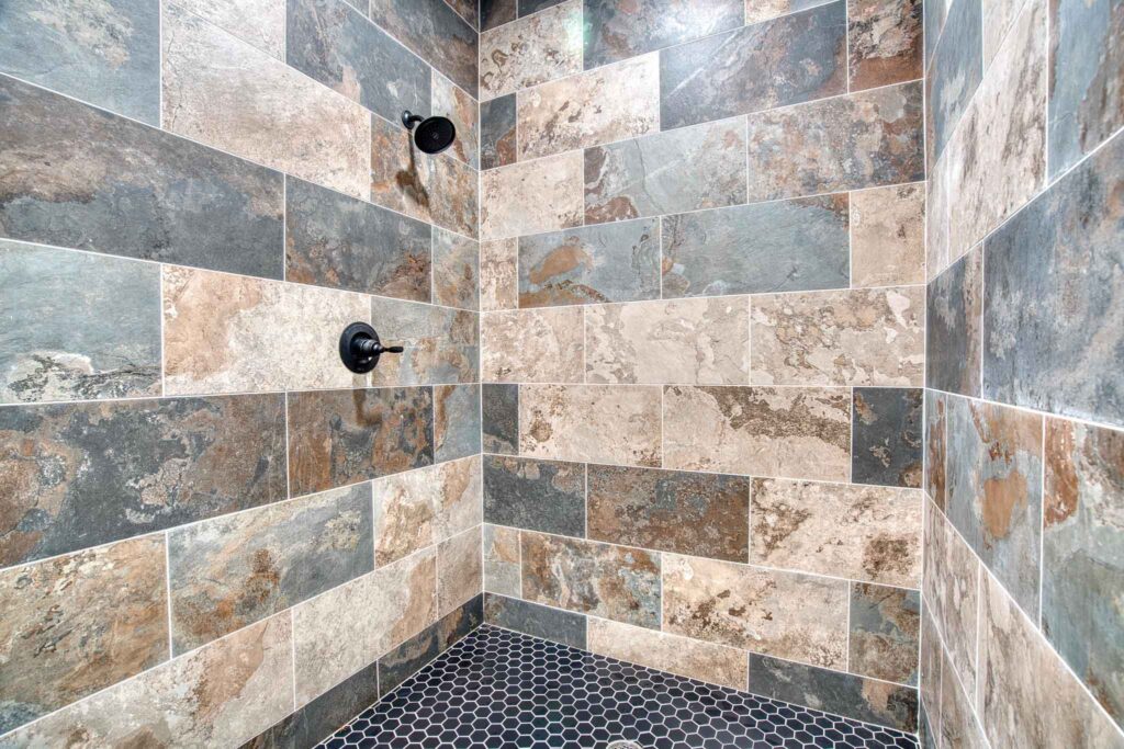A modern shower with walls of large, rectangular slate tiles in earthy tones is reminiscent of the serene landscapes surrounding Lake Tenkiller Camping. The floor boasts small hexagonal black tiles, and two black shower fixtures are elegantly mounted on the wall.